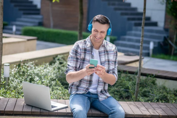 Fröhlicher dunkelhaariger Mann mit Kopfhörer, Smartphone checkend, mit Laptop auf Bank sitzend — Stockfoto