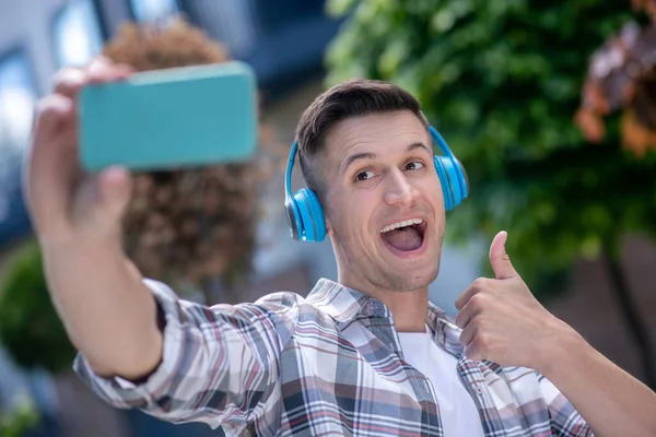 Alegre macho moreno usando auriculares, haciendo selfie, sonriendo, pulgar hacia arriba — Foto de Stock