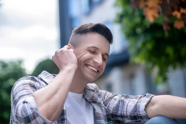 Cheerful dark-haired male wearing wireless earphones, smiling, listening to music, touching his ear — Stock Photo, Image