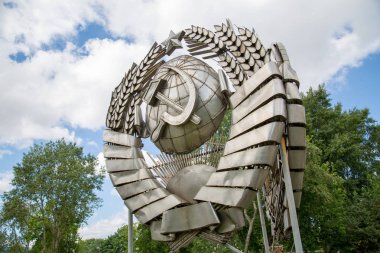 Mavi gökyüzü ağaçlarının arka planında paslanmaz çelikten yapılmış SSCB arması. Geleneğin sembolleri.