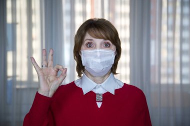 A middle- aged woman in a white medical mask with a raised hand and fingers showing the word OK in a room of an apartment building , protection from viral infection, flu, tuberculosis. Medicine, protection from SARS -CoV-2 coronavirus clipart