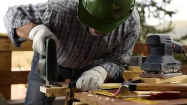 Kasklı, deri eldivenli, elektrikli testereli, tahta bir masa kesmeye çalışan yetişkin bir marangoz. İnşaat endüstrisi, ev işi, kendin yap. Görüntü.