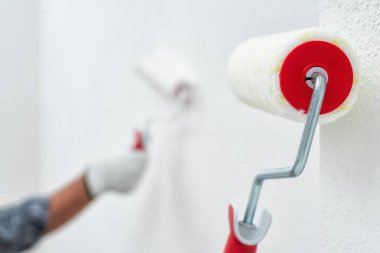 Beyaz tulumlu, koruyucu eldivenli, beyaz boyalı bir ev boyacısı. Duvarı silindir kullanarak beyaz boyayla boyuyor. İnşaat endüstrisi. İş güvenliği.