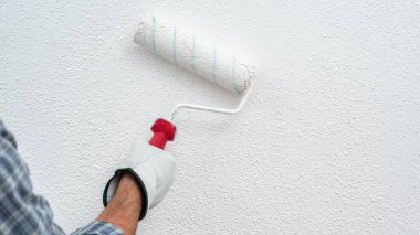Beyaz tulumlu, koruyucu eldivenli, beyaz boyalı bir ev boyacısı. Duvarı silindir kullanarak beyaz boyayla boyuyor. İnşaat endüstrisi. İş güvenliği.