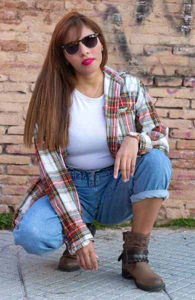 Peruvian girl with sunglasses on a sunset with brick wall in the background, checkered t-shirt and jeans.