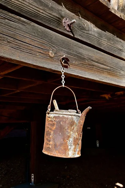 A very old rusty can with a spout for pouring liquids hangs from a chain attached to a beam of an old mining structure.