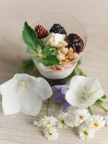 Greek yogurt with blackberry jam and nuts, mint and blackberries on top, with flowers around on wooden surface