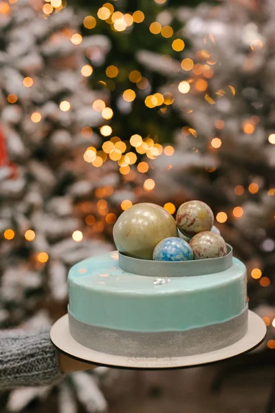View of  Christmas cake with chocolate balls on top on female hand with blurred lights on background
