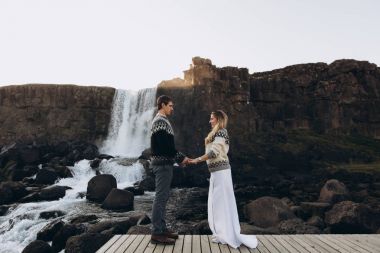 Şelale arka plan üzerinde el ele tutuşarak kazak örme sıcak genç çift giymiş