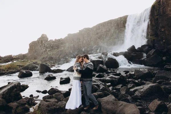 Young couple dressed in knitted jumpers cuddling on waterfall background