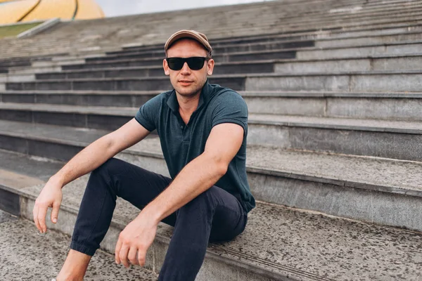 View of cool dude wearing dark glasses, dark jeans and t -shirt sitting on stairs