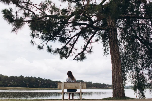 Nehir ve orman arka plan üzerinde büyük ağaç yakınındaki bankta oturan kadın arkadan görünüş