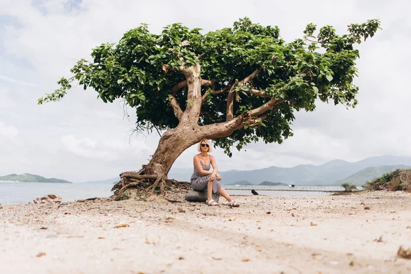 木の近くの海岸に座っている縞模様のドレスを着ている女性のビュー — ストック写真