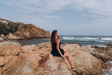 Cliff üzerinde oturan küçük siyah elbise giymiş esmer genç kadın yan görünüm.