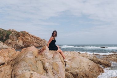 Cliff üzerinde oturan küçük siyah elbise giymiş esmer genç kadın yan görünüm.