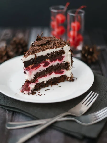 巧克力蛋糕与樱桃和酸奶馅的看法在白色盘子服务在黑木桌背景的叉子 餐巾和松树锥体 — 图库照片
