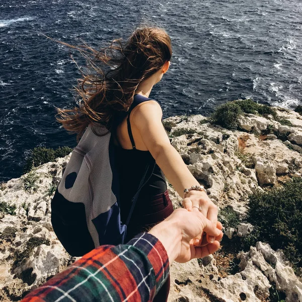 風の強い日で暗い水と海岸でロッキー山脈の上に男性の手を握ってのバックパックと若い女性の背面します — ストック写真
