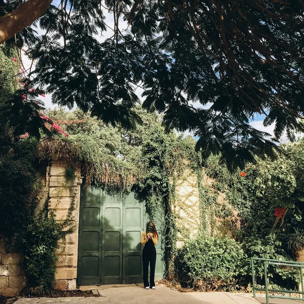 Woman wearing black romper, yellow jumper and white sneakers standing near green door in old garden.