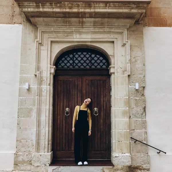 Woman wearing black romper, yellow jumper and white sneakers standing near house entrance and bowed head.