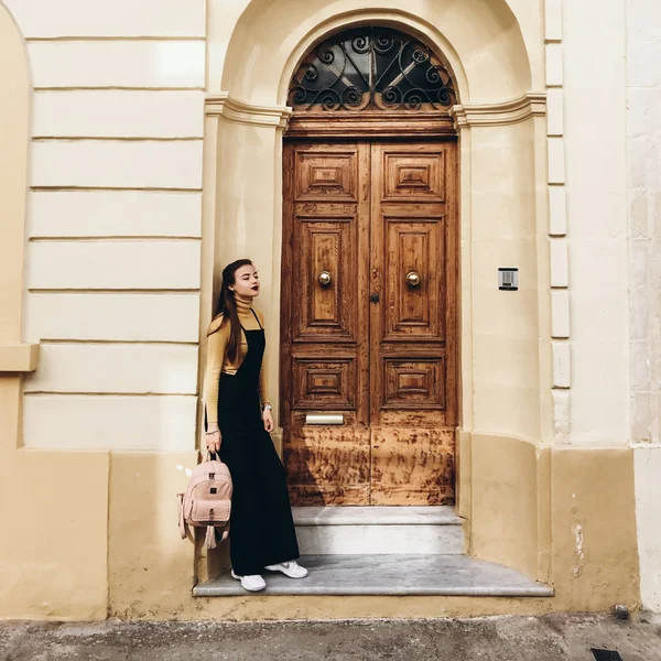 Woman wearing black romper, yellow jumper and white sneakers standing near house entrance and holding pink backpack in hand.