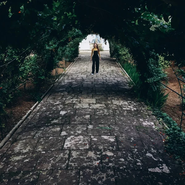 Woman wearing black romper, yellow jumper and white sneakers having fun and walking on alley in old garden.