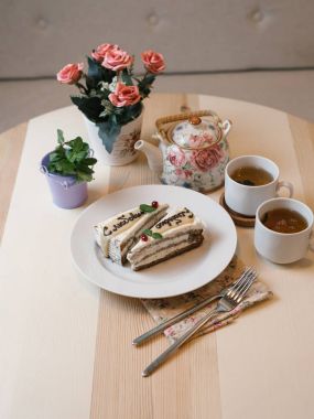 lezzetli dilim kek beyaz plaka üzerinde metin ile yakın iki beyaz fincan çay çaydanlık, çatal, peçete ve ahşap masa arka plan üzerinde çiçekler ile çekim