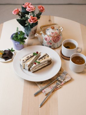 lezzetli dilim kek beyaz plaka üzerinde metin ile yakın iki beyaz fincan çay çaydanlık, çatal, peçete ve ahşap masa arka plan üzerinde çiçekler ile çekim