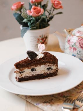 Çikolatalı kek krem peynir dolum ile oreo kurabiyesinin ile dekore edilmiş ve beyaz tabakta çatal, peçete, fincan çay, çaydanlık ve çiçek buketi ile krem şanti