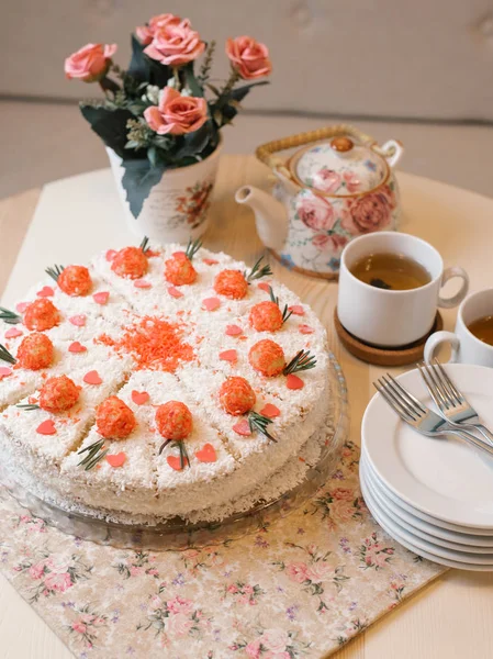 Sponge cake with tender cream, berries filling, coconut chips and decorative sweet flowers on top served with teapot, cup of tea, plates and forks