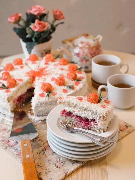 Sponge cake with tender cream, berries filling, coconut chips and decorative sweet flowers on top served with tea set, forks,  spatula and flower bouquet on background