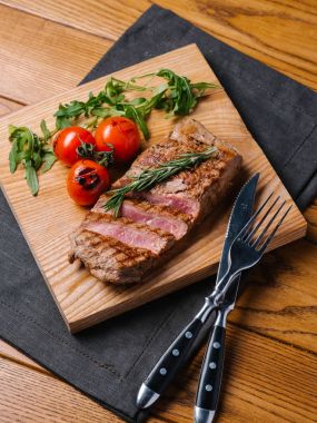 Bıçak ve çatal ahşap kesme tahtası ve gri peçete ile iştah açıcı orta nadir sığır eti biftek ızgara domates, biberiye ve roka ile servis
