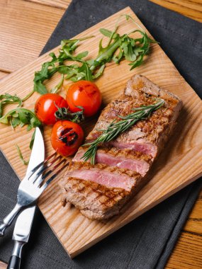 Bıçak ve çatal ahşap kesme tahtası ve gri peçete ile iştah açıcı orta nadir sığır eti biftek ızgara domates, biberiye ve roka ile servis