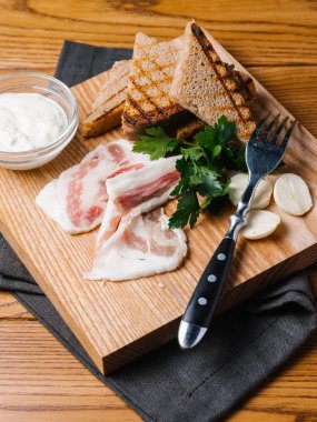 Dilimlenmiş domuz pastırması, sarımsak dilimleri ve maydanoz, Sarımsak soslu ızgara ekmek tost ahşap kesme tahtası çatal ve gri peçete ile servis