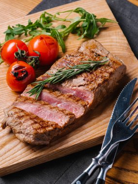 Bıçak ve çatal ahşap kesme tahtası ve gri peçete ile Görünümü Kapat orta nadir sığır biftek ızgara domates, biberiye ve roka ile servis