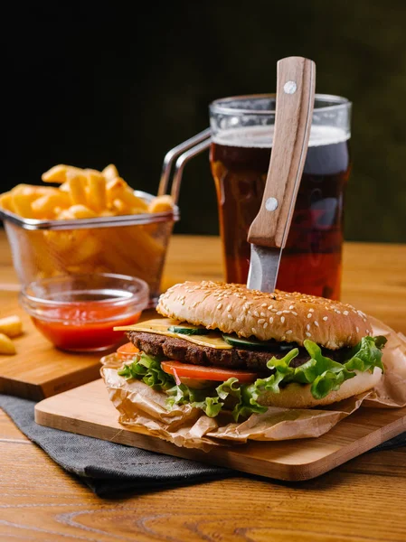 Fresh cooked cheeseburger with knife inside, french fries in basket, cold dark beer and sweet ketchup served on wooden chopping boards and grey napkin