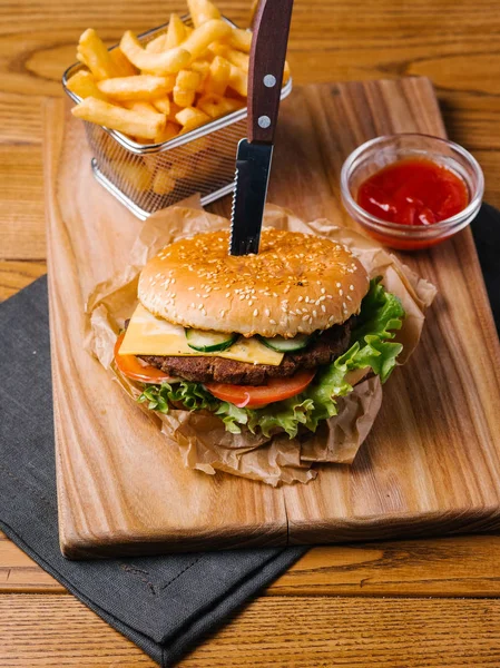 Cheeseburger with knife inside, french fries in basket and sweet ketchup served on wooden chopping board