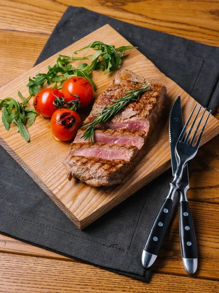 Bıçak ve çatal ahşap kesme tahtası ve gri peçete ile iştah açıcı orta nadir sığır eti biftek ızgara domates, biberiye ve roka ile servis