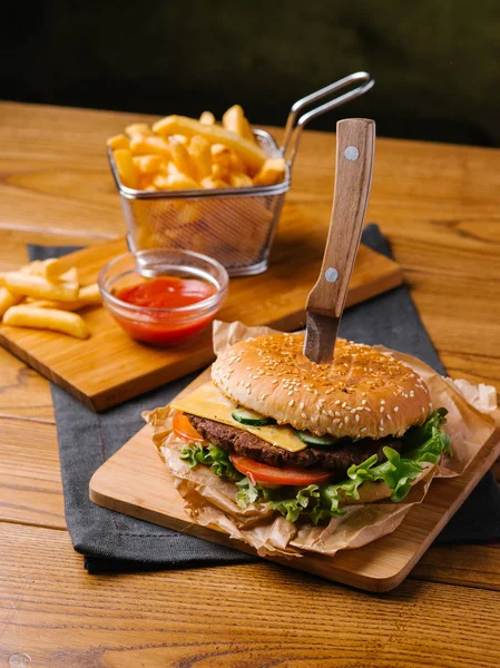 Fresh cooked cheeseburger with knife inside, french fries in basket and sweet ketchup served on wooden chopping boards and grey napkin