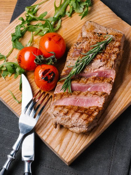Bıçak ve çatal ahşap kesme tahtası ve gri peçete ile Görünümü Kapat orta nadir sığır biftek ızgara domates, biberiye ve roka ile servis