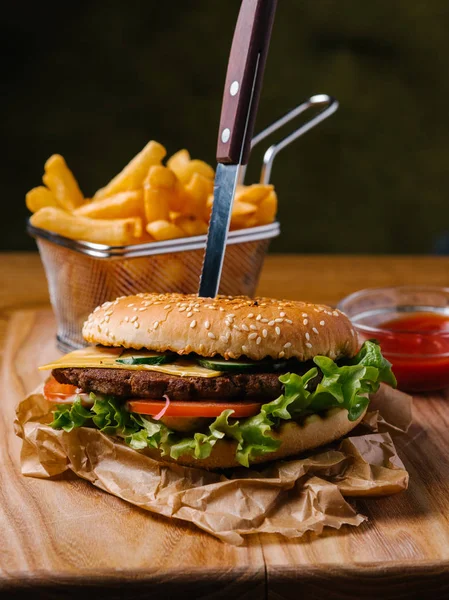 Cheeseburger with knife inside, french fries in basket and sweet ketchup served on wooden chopping board