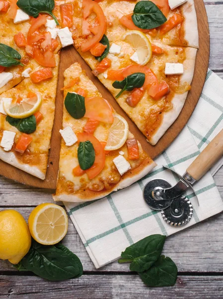 Pizza with salmon, feta, basil leaves, tomatoes, lemon slices and mozzarella served on pizza plate with pizza cutter and checkered napkin