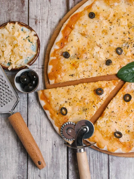 Top view of pizza with mozzarella, parmesan, tomato sauce and olives served on wooden pizza plate with pizza cutter, grater on shabby wooden planks background