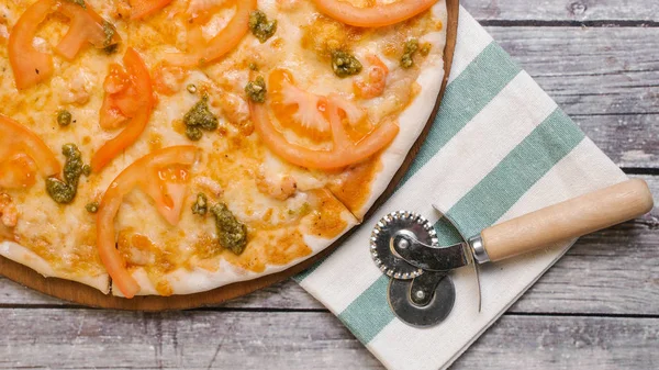 Pizza with tomato sauce, pesto sauce, mozzarella and tomatoes served on wooden pizza plate with pizza cutter and striped napkin