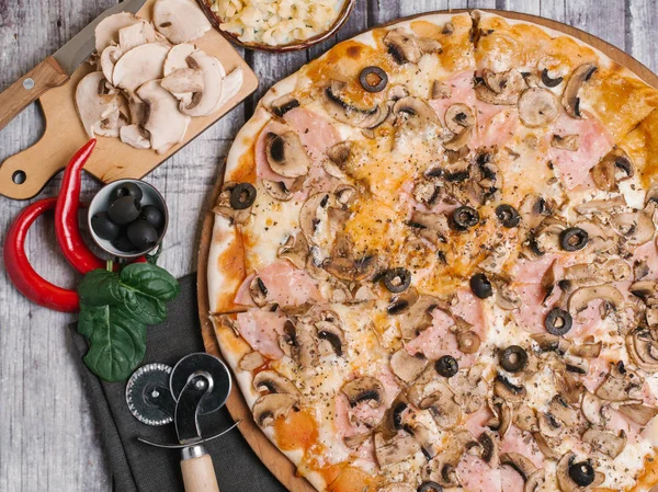 Pizza with tomato sauce, mozzarella, ham, olives and mushrooms served on wooden pizza plate with napkin, pizza cutter, chilli peppers, shinach leaves, knife and sliced mushrooms on chopping board on wooden planks background