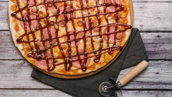 Pizza with tomato sauce, barbeque sauce, mozzarella, sausages served on wooden pizza plate with pizza cutter and napkin