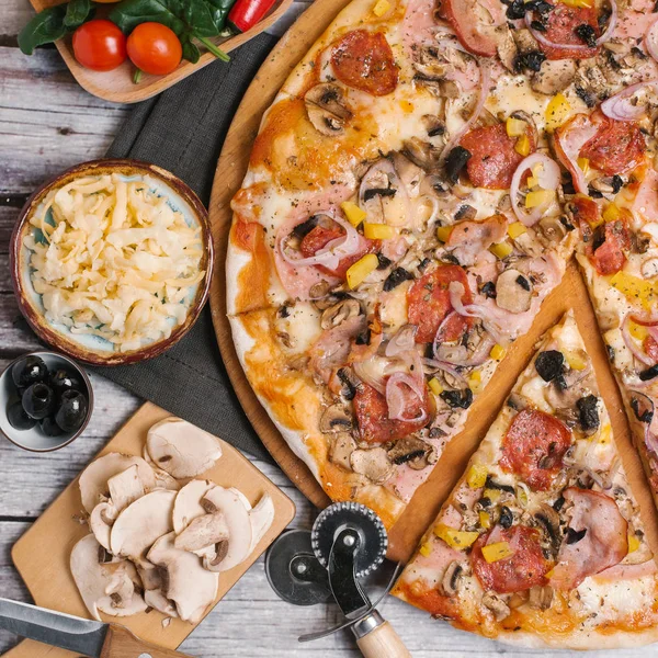 Pizza with tomato sauce, mozzarella, ham, sausages, olives, sweet onions, yellow peppers and mushrooms served on wooden pizza plate with pizza cutter, napkin and veggies on wooden planks background