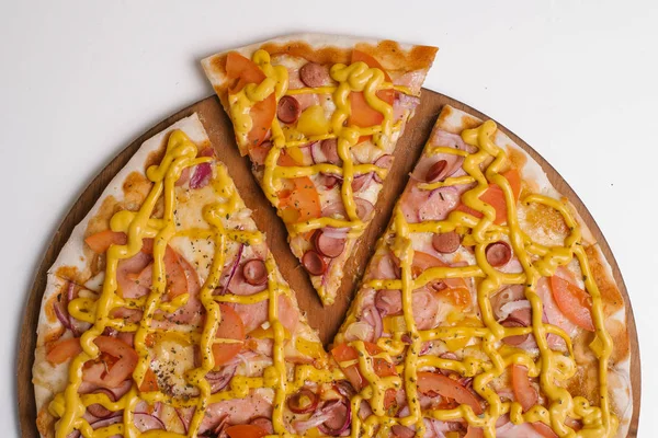 Pizza with tomato sauce, mustard sauce, mozzarella, ham, sausages, tomatoes, yellow peppers and sweet onions served on wooden pizza plate on white table
