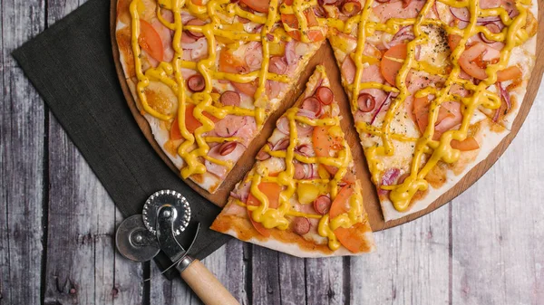 Pizza with tomato sauce, mustard sauce, mozzarella, ham, sausages, tomatoes, yellow peppers and sweet onions served on wooden pizza plate with pizza cutter and napkin on shabby planks background