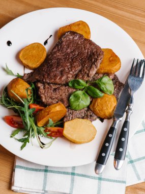 Fırında patates, taze domates, roka, ıspanak ile aromatik sığır eti dilim beyaz bıçak ve çatal ile tabakta