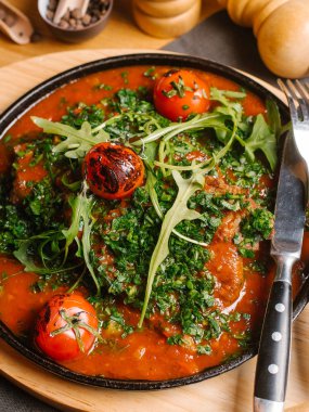 Domates soslu et pişmiş, taze kiraz domates, roka ve doğranmış yeşil ahşap kesme tahtası peçete, çatal ve bıçak ile tava servis